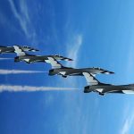 Four military jets flying in formation against a blue sky leaving smoke trails