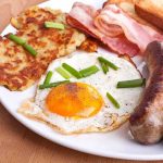 A hearty breakfast plate featuring a fried egg, crispy bacon, sausages, hash browns, and toasted bread garnished with green onions