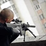 Person aiming a rifle from a building balcony.