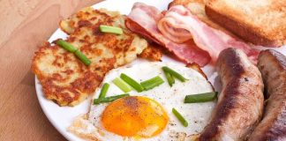 A hearty breakfast plate featuring a fried egg, crispy bacon, sausages, hash browns, and toasted bread garnished with green onions