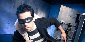 A masked thief in a striped shirt holding a bag while emerging from a bank vault