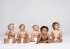 Five diverse babies sitting on a neutral background, displaying playful expressions
