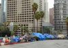 One-Way Bus Tickets in 1996. Trash Trucks in 2026. Same City. Row of tents along a city street with skyscrapers in the background