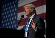 man in suit speaking at a podium with a US flag backdrop