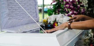 Autopsy Hold Freezes Painful Truth hands resting on a white casket with flowers nearby