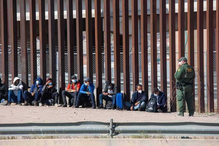 Group of people sitting by a tall border fence with an officer standing nearby