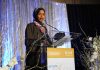Federal Raid Hits Omar’s Home Ilhan Omar speaking at a podium on a decorated stage