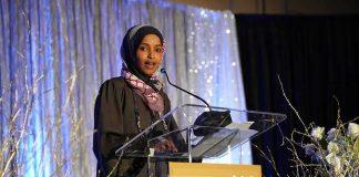 Federal Raid Hits Omar’s Home Ilhan Omar speaking at a podium on a decorated stage
